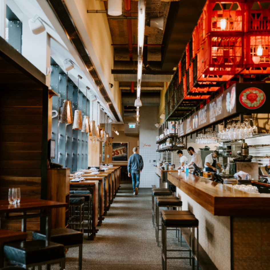 Restaurant interior with a long bar, high tables, chefs working, and a person walking towards the back.