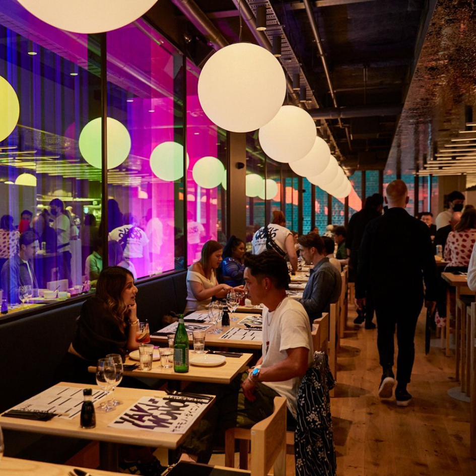 Busy restaurant interior with round pendant lights, colorful window panels, and diners eating meals at wooden tables.