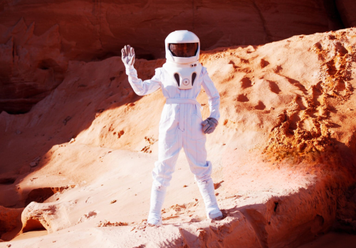 A person waving, dressed as an astronaut in a barren landscape.